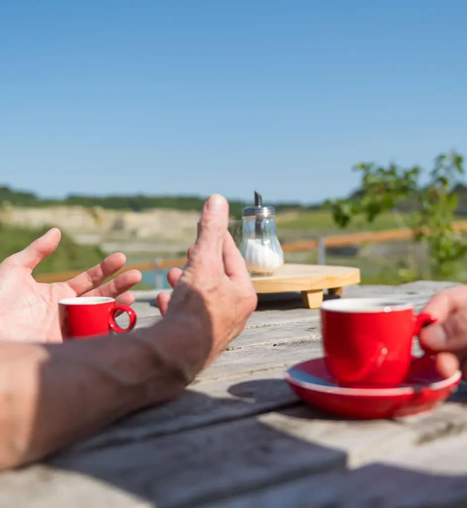 Handen van twee mensen in gesprek aan een houten tafel met rode koffiekopjes